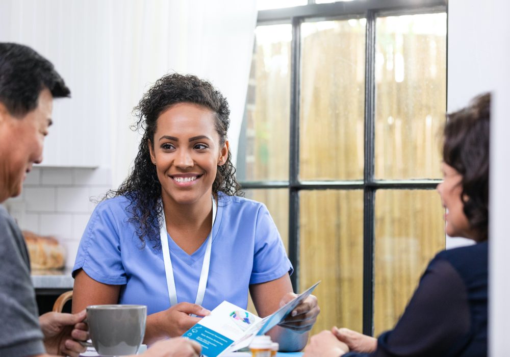 The senior adult man and his wife smile as he takes the brochure the visiting nurse holds out to him.