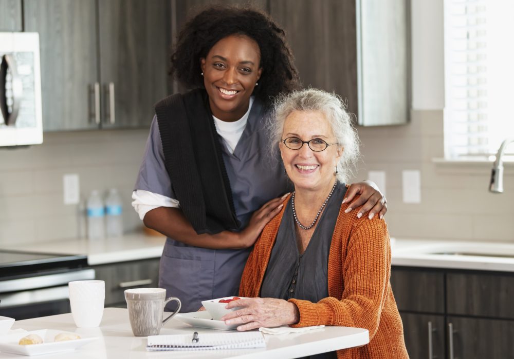 A senior woman in her 80s sitting at home in her kitchen. A home caregiver is standing beside her and they are smiling at the camera. The healthcare worker is an African-American woman in her 30s.